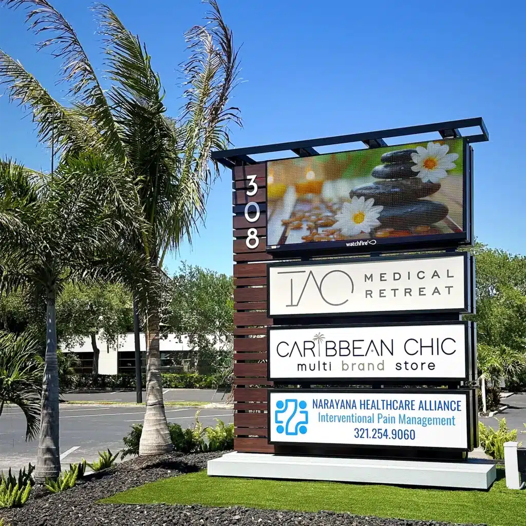 A freestanding sign for multiple businesses is shown. The top section features a 10MM full color electronic message center sign by Watchfire, showcasing an image of spa stones and flowers. Below, there are signs for various businesses: TAO Medical Retreat, Caribbean Chic multi-brand store, and Narayana Healthcare Alliance for Interventional Pain Management with a phone number 321.254.9060. The sign is located at 308 and is surrounded by palm trees and a landscaped area.