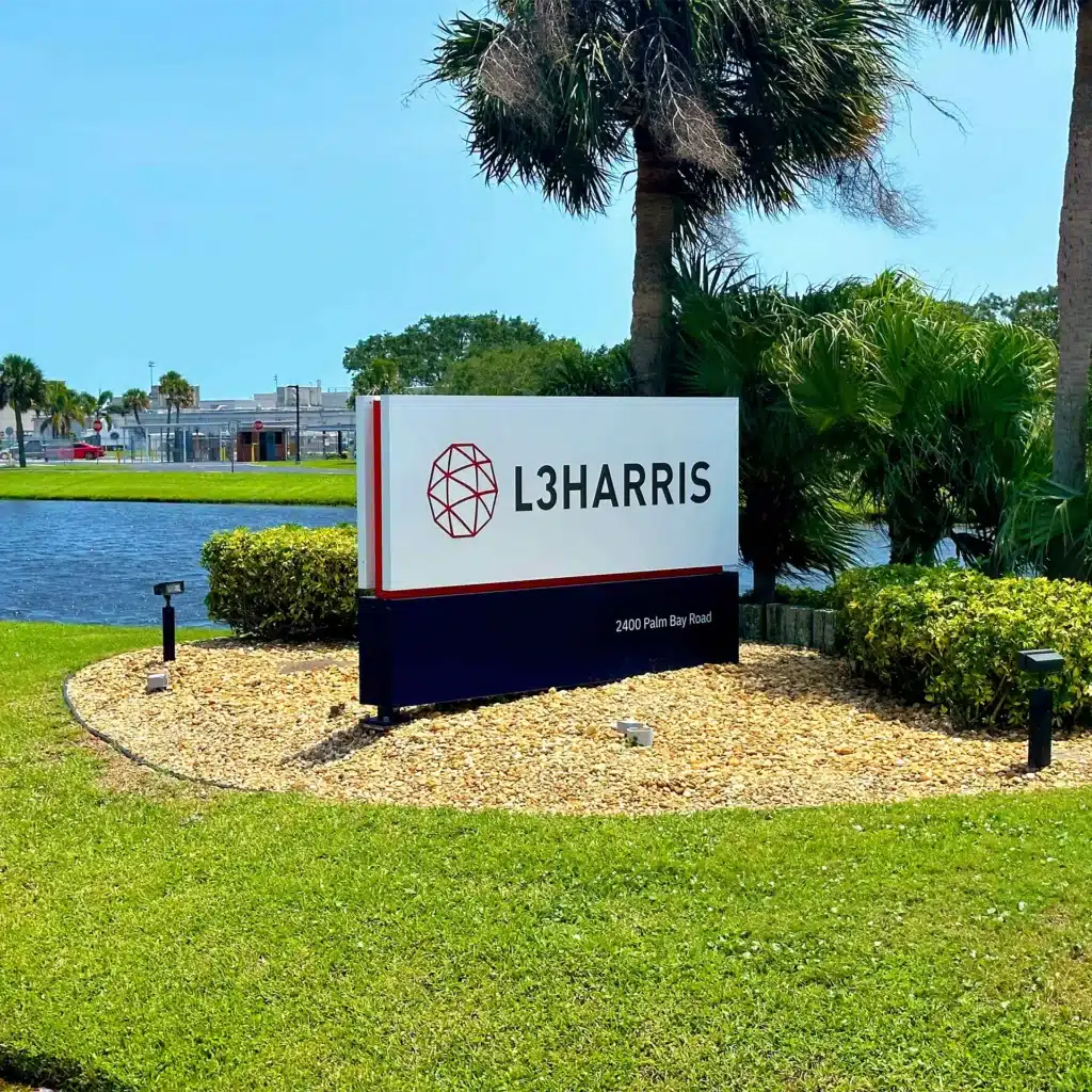 L3Harris low profile monument sign at 2400 Palm Bay Road, surrounded by lush greenery and palm trees, with a pond in the background under a clear blue sky.