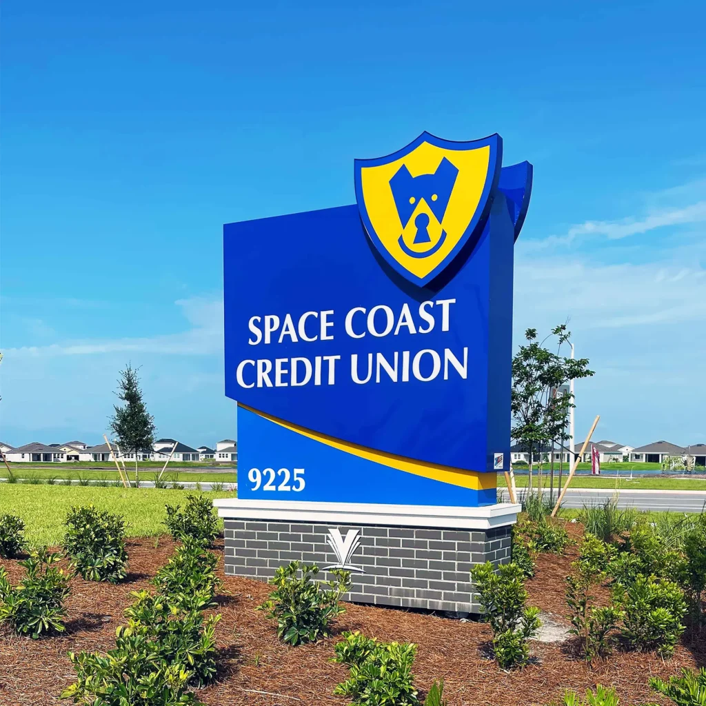 Space Coast Credit Union blue and yellow low profile monument sign at Viera Branch with logo and address 9225, surrounded by landscaped greenery under a clear blue sky.